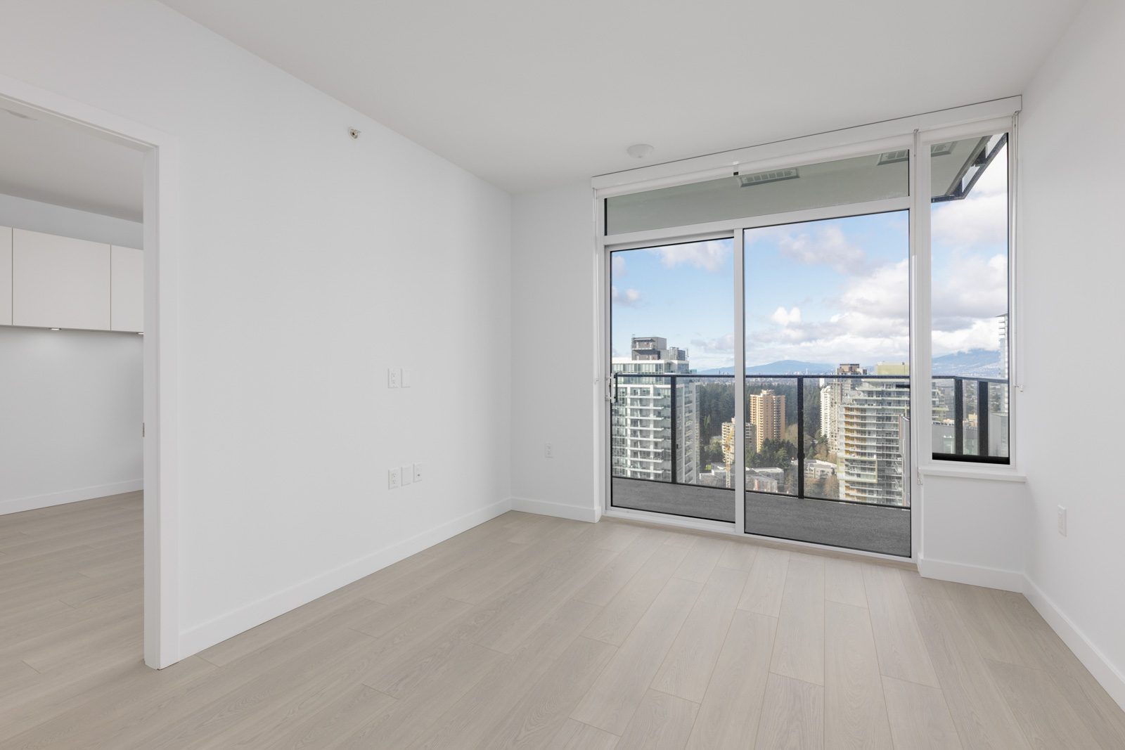 Unfurnished modern apartment room with light wood flooring, white walls, large window doors leading to a balcony, and a city skyline view.