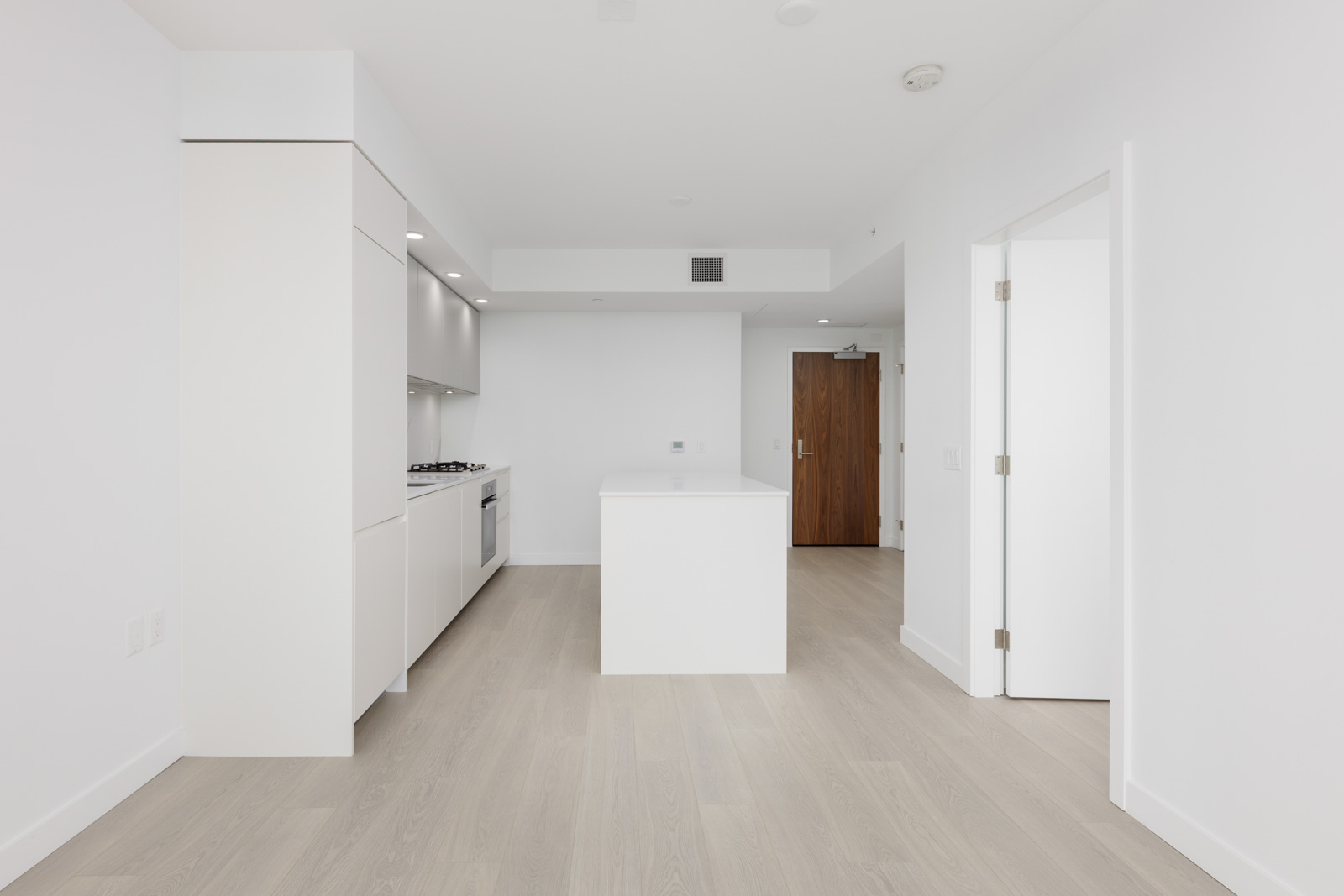 A modern, minimalistic kitchen with white cabinets, a center island, light wood flooring, and an open doorway leading to another room.