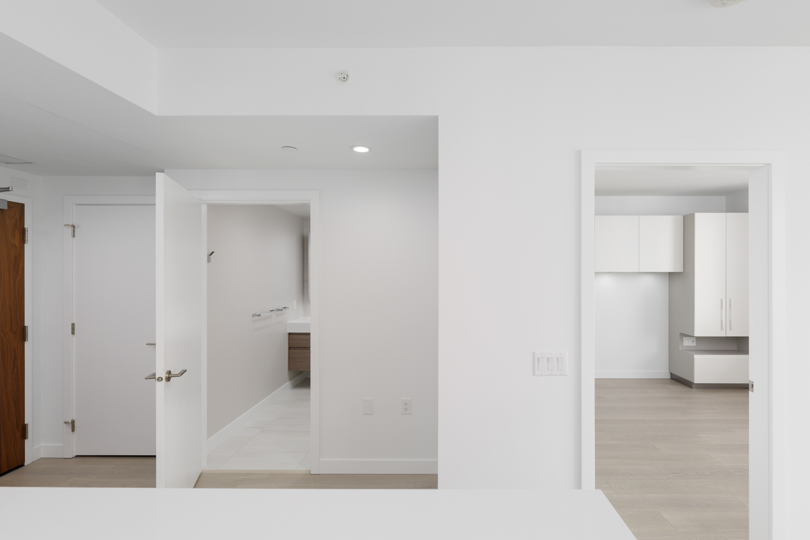 Modern, unfurnished apartment interior with white walls, open doors revealing a bathroom and another room, and light wood flooring.
