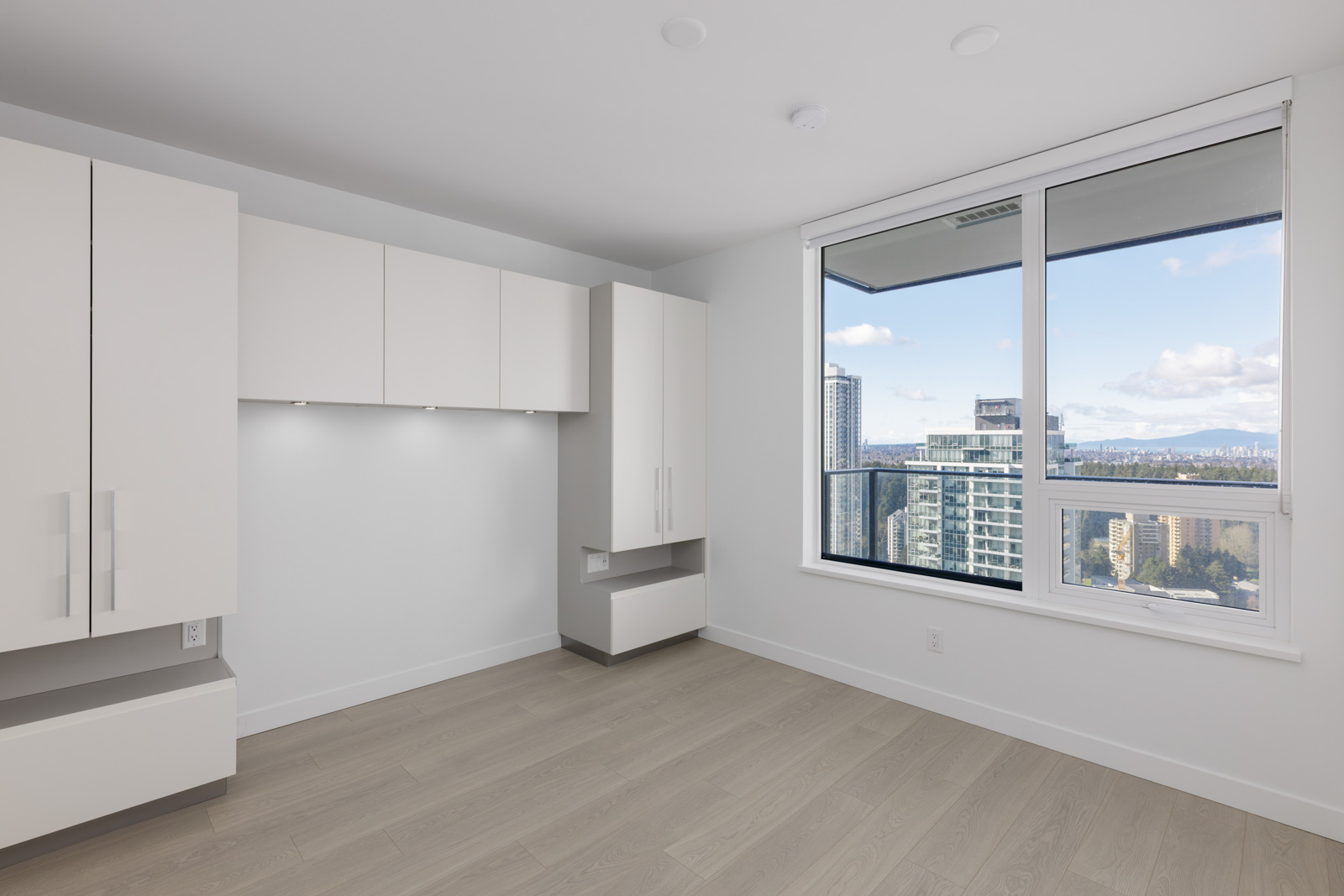 Modern, unfurnished apartment room with built-in white cabinets, light wood flooring, and a large window overlooking city buildings and a distant skyline.