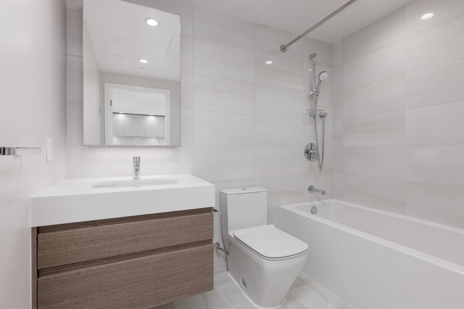 Modern bathroom with a wall-mounted sink, rectangular mirror, toilet, and bathtub with a showerhead. The design features light-colored tiles and wooden cabinetry.