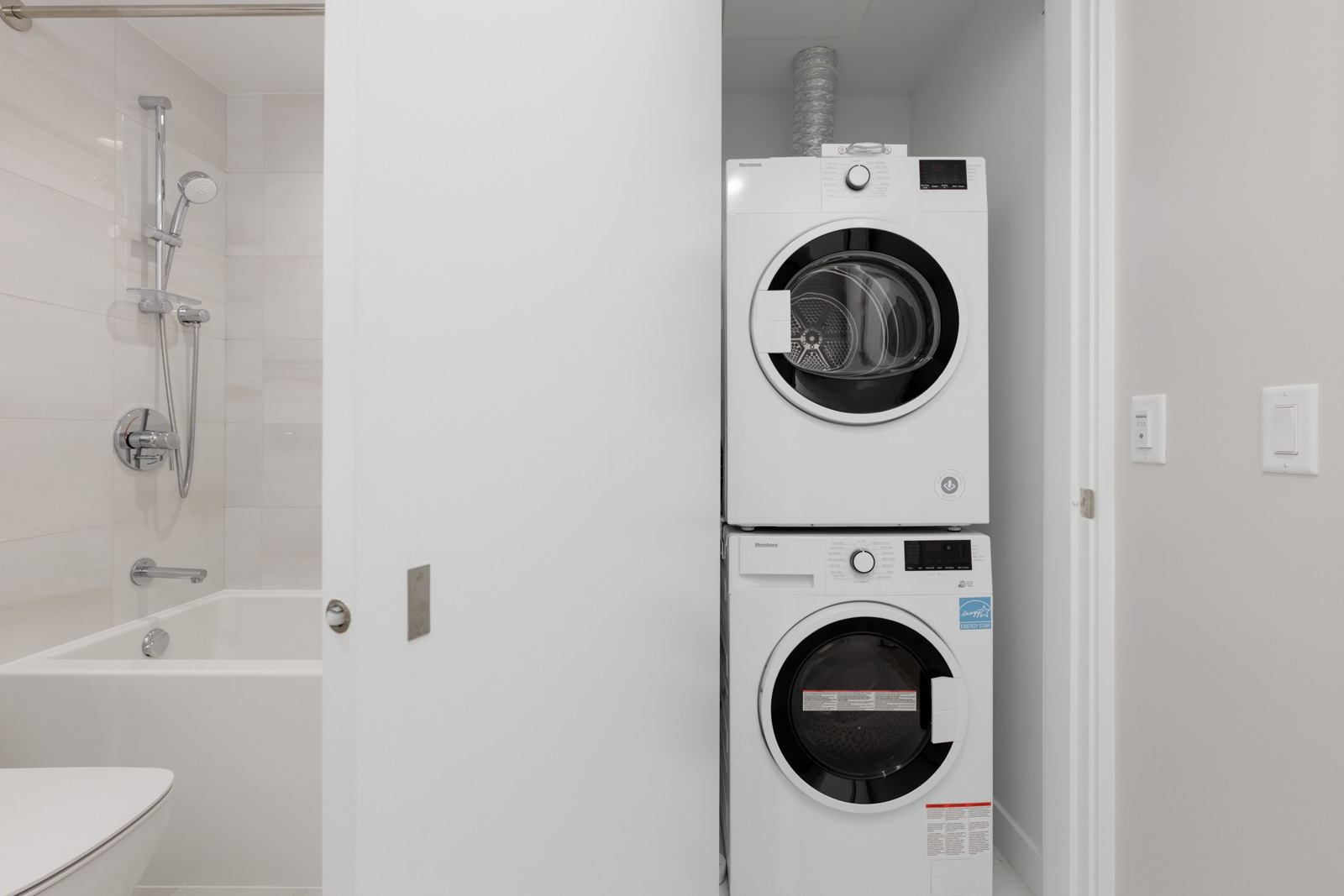 A modern bathroom with a bathtub and shower next to a stacked washer and dryer unit in a white alcove.