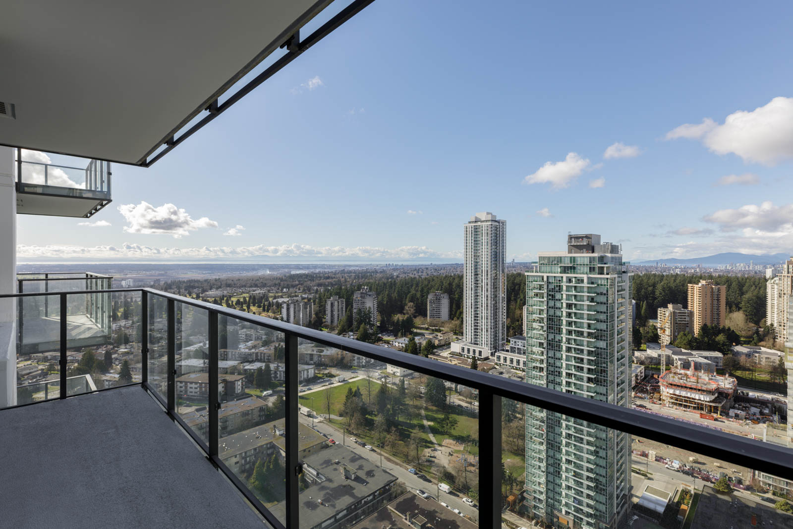 View from a high-rise balcony overlooking a cityscape with tall buildings, roads, trees, and distant mountains under a partly cloudy sky.