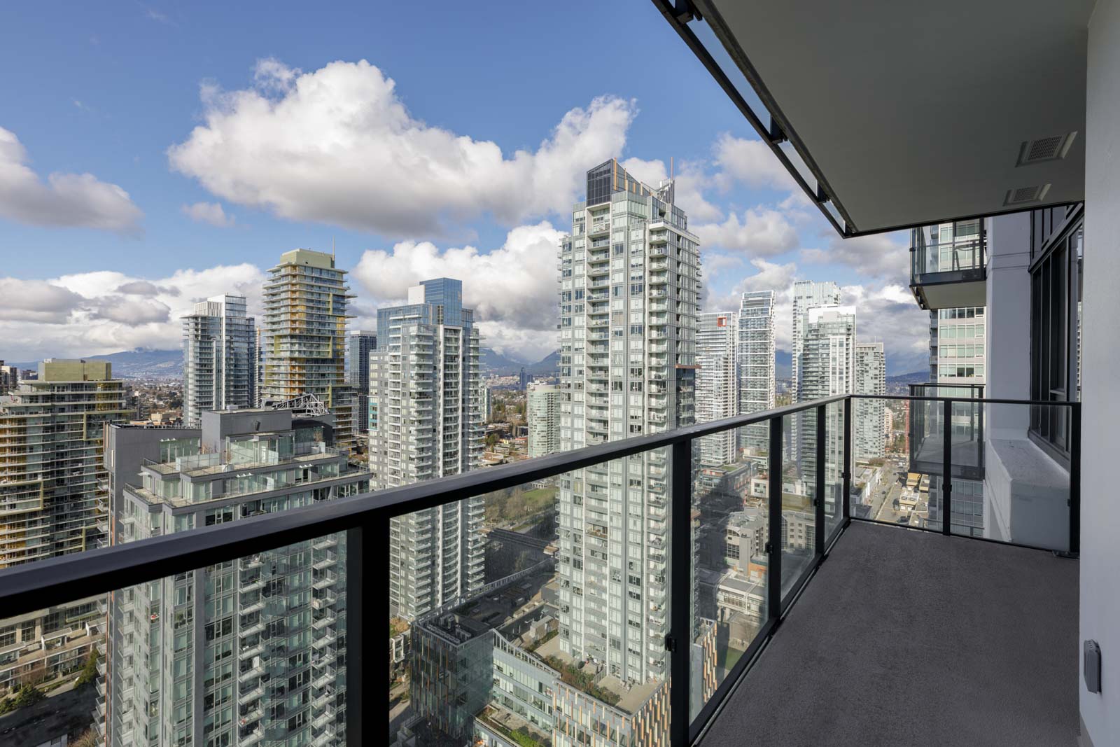 High-rise cityscape view from a balcony with glass railing, showing multiple modern buildings under a partly cloudy sky.