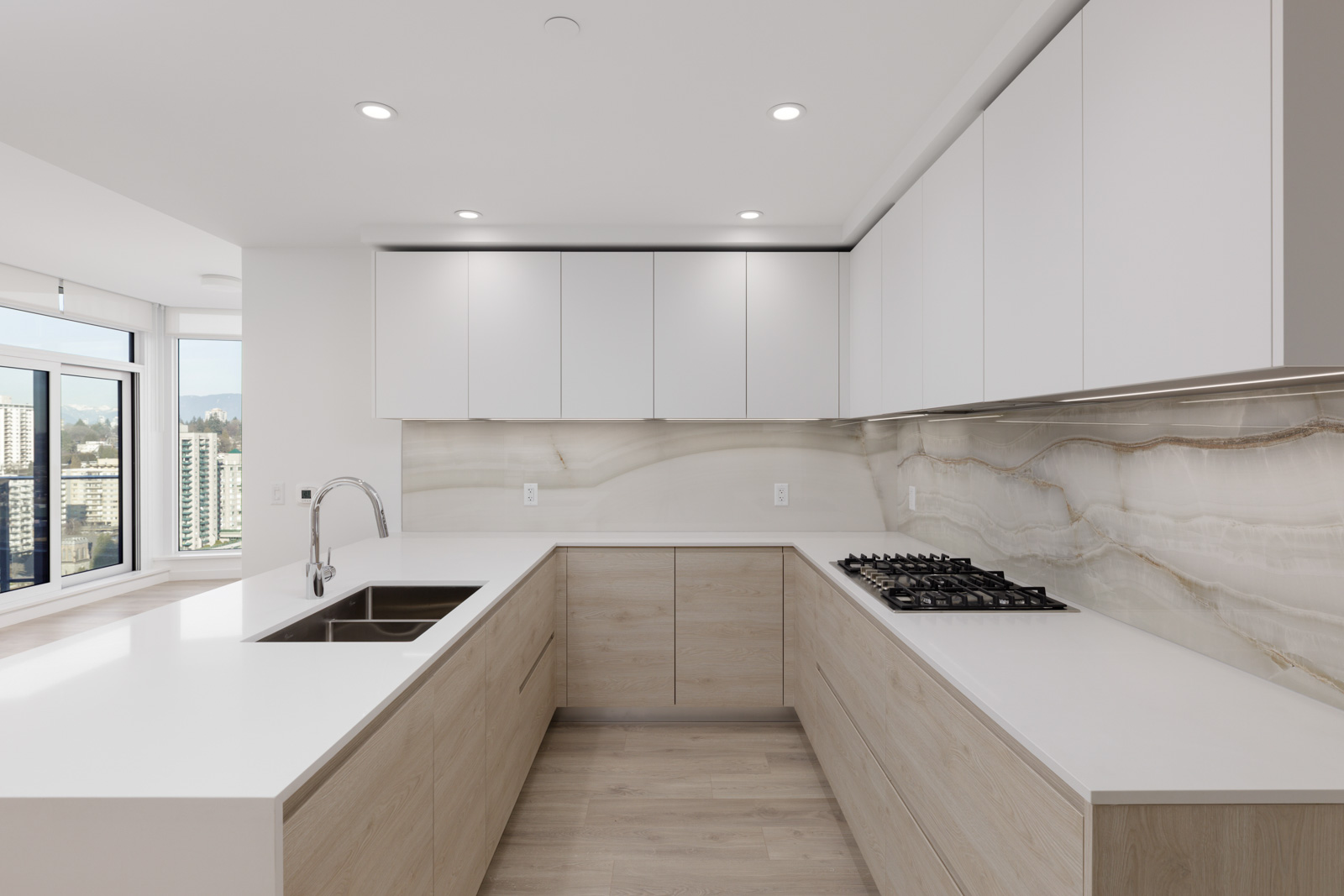 Modern kitchen with light wood cabinets, white countertops, stainless steel sink, gas cooktop, and large windows with a city view.