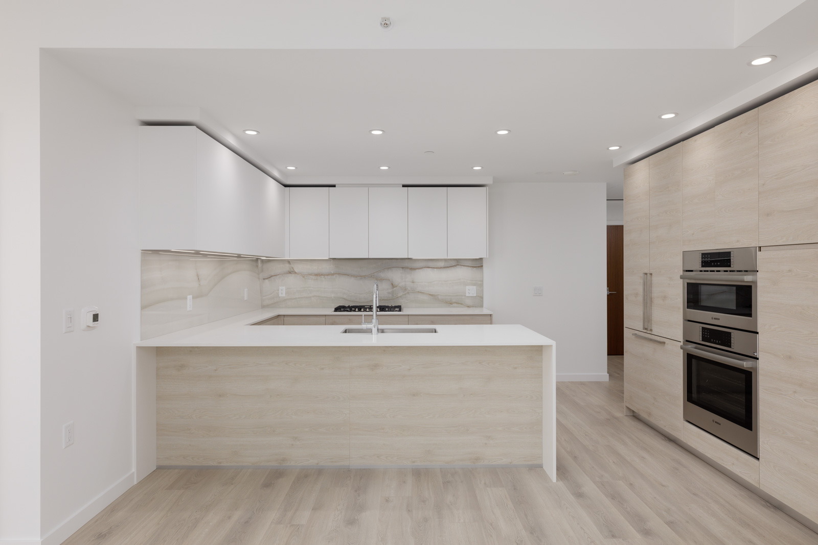 Modern kitchen with light wood cabinets, white countertops, built-in oven, stainless steel appliances, and recessed lighting.