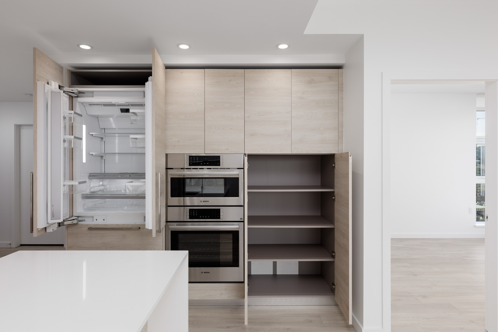 Modern kitchen with light wood cabinets, built-in appliances, an open refrigerator, and open pantry shelves; white walls and floors, minimalistic design.