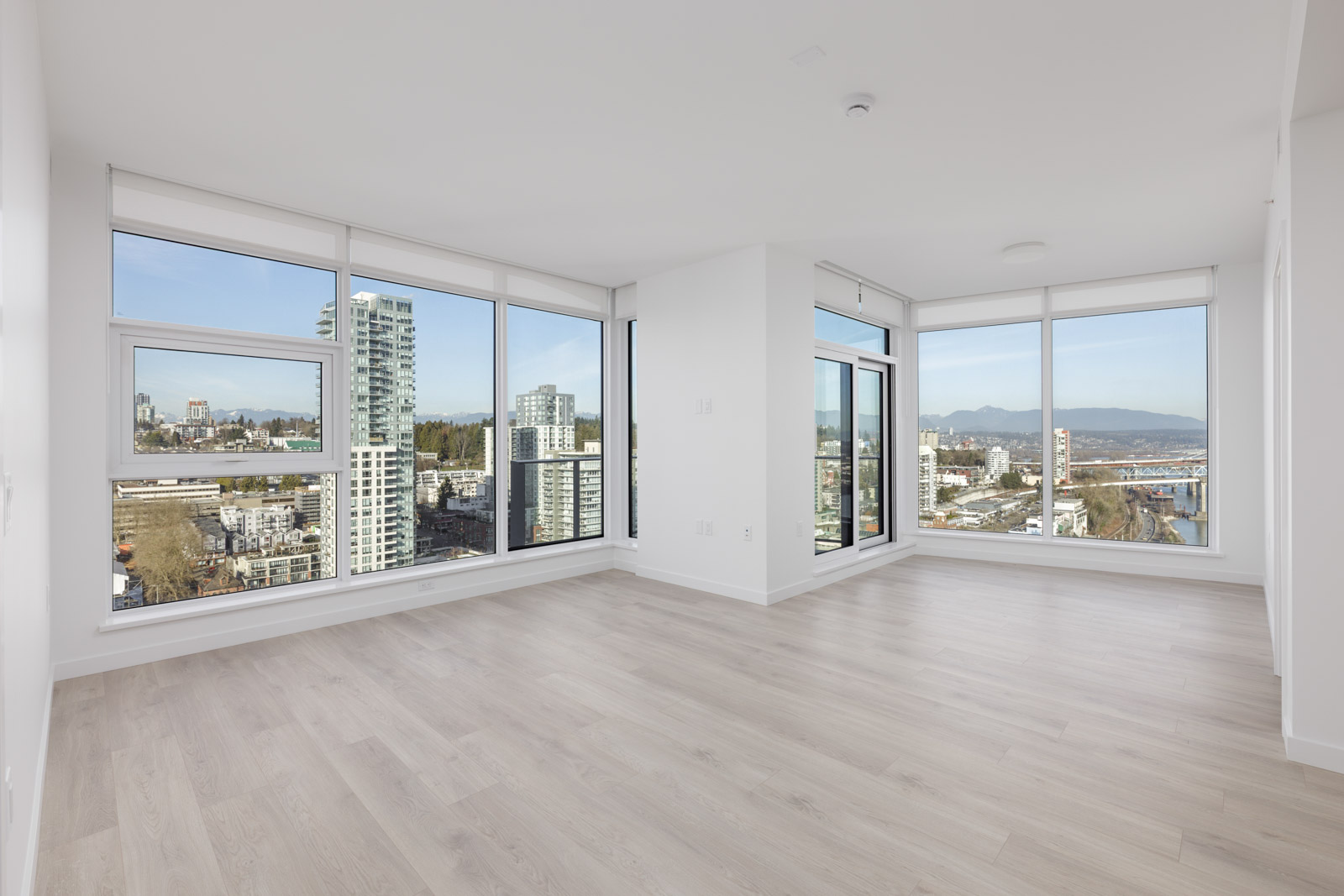 A modern, empty apartment with large floor-to-ceiling windows, light wood floors, and city views of high-rise buildings and mountains.