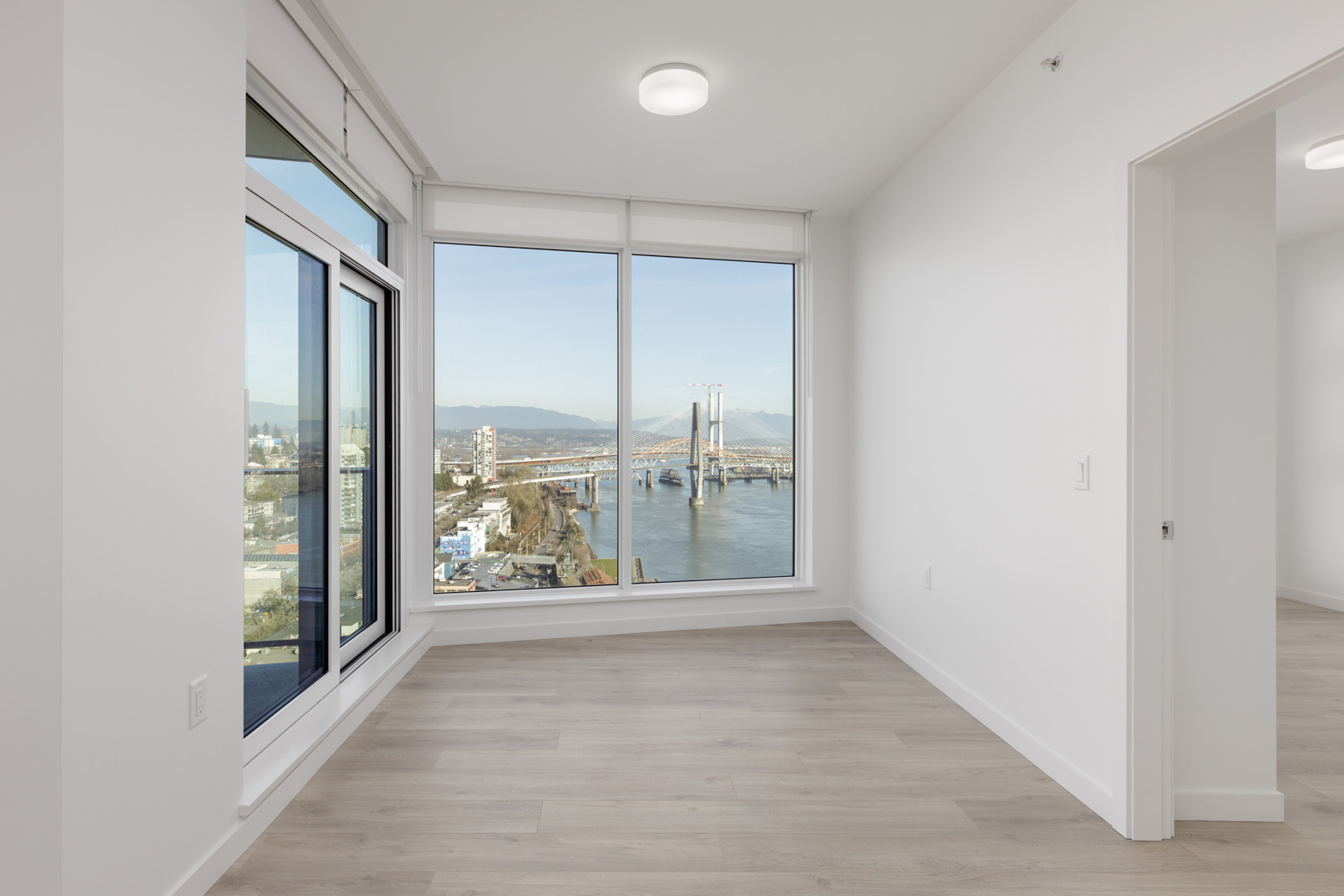 Modern empty room with large windows, light wood floors, and a view of a cityscape, river, and bridge.