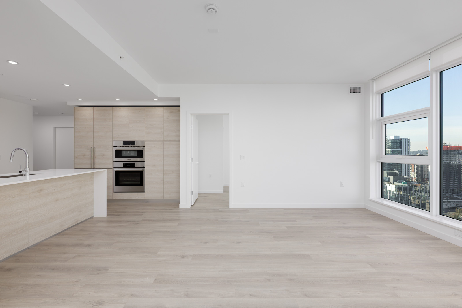 A modern, unfurnished apartment with light wood floors, large windows with a city view, and a kitchen area featuring built-in appliances and an island.