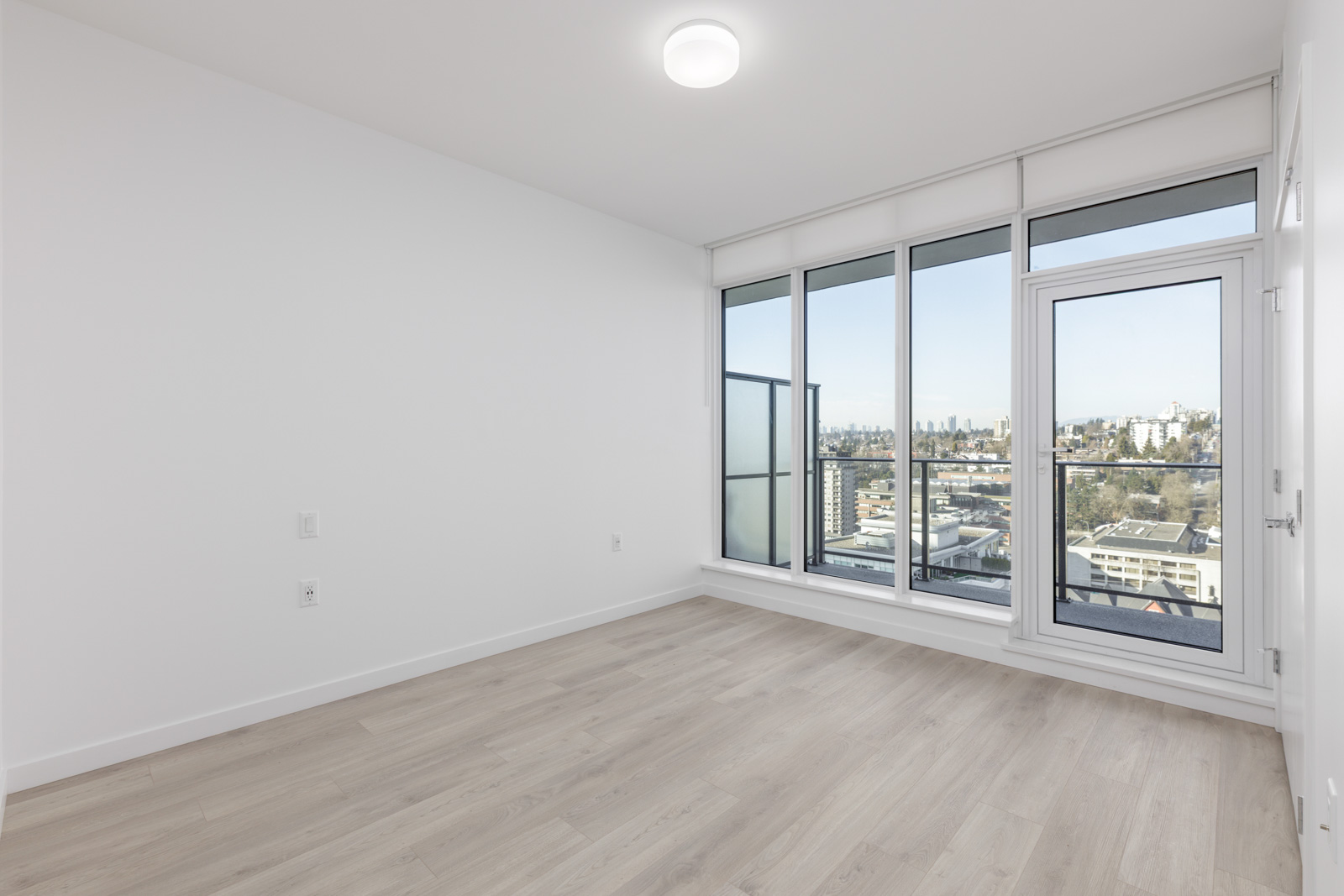 Empty modern room with light wood flooring, white walls, and large floor-to-ceiling windows leading to a balcony with a cityscape view.