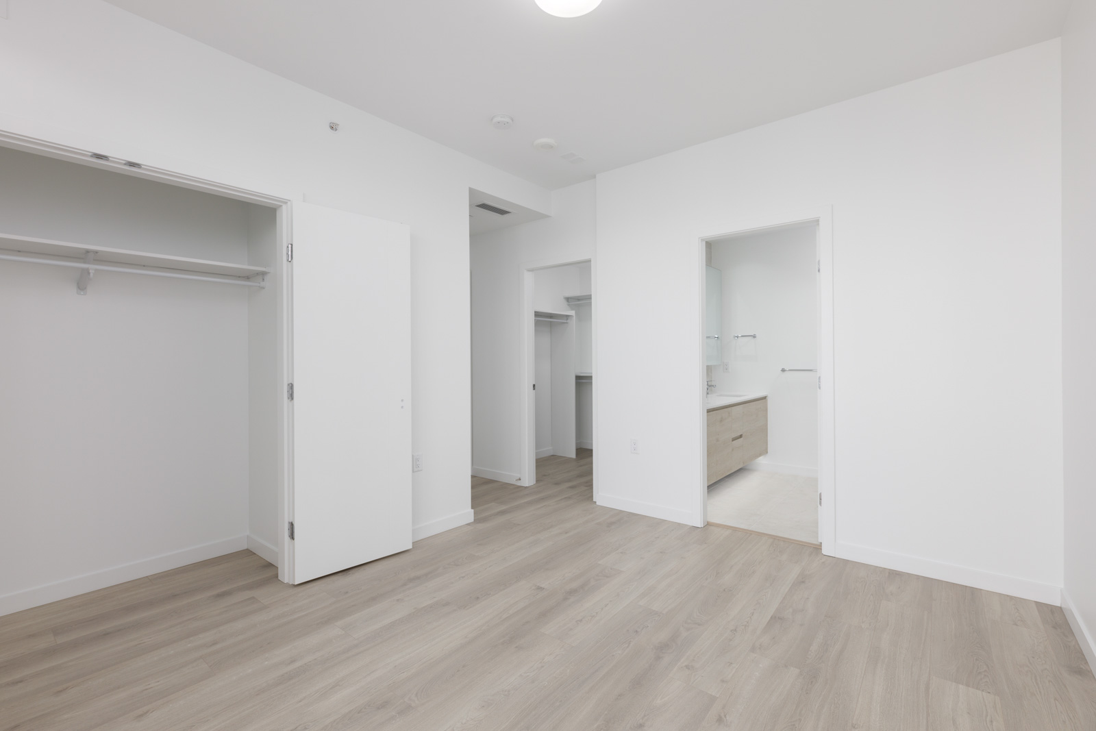Empty room with light wood flooring, open closet, white walls, and an open door leading to a bathroom with a double sink vanity.