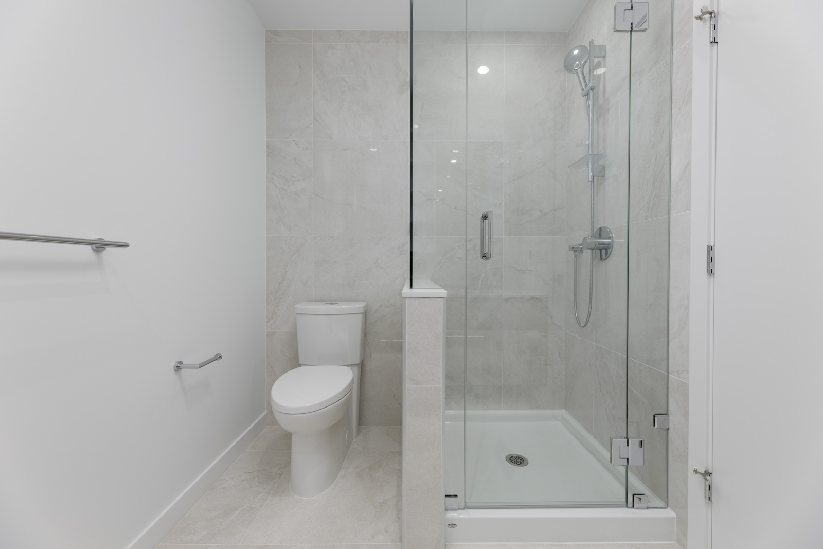 Modern bathroom with a white toilet next to a glass-enclosed shower. The walls and floor are tiled in light gray, and a metal towel bar is mounted on the wall.