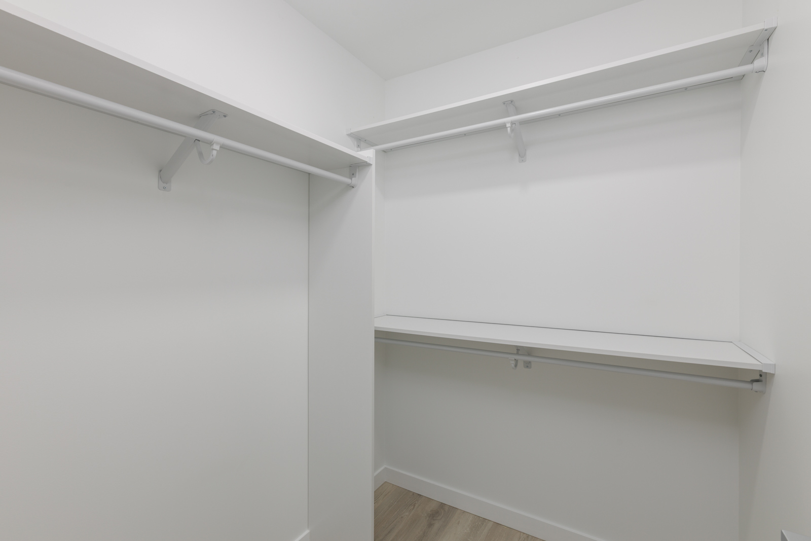 Empty walk-in closet with white walls, built-in white shelves, and hanging rods, featuring light wood flooring.
