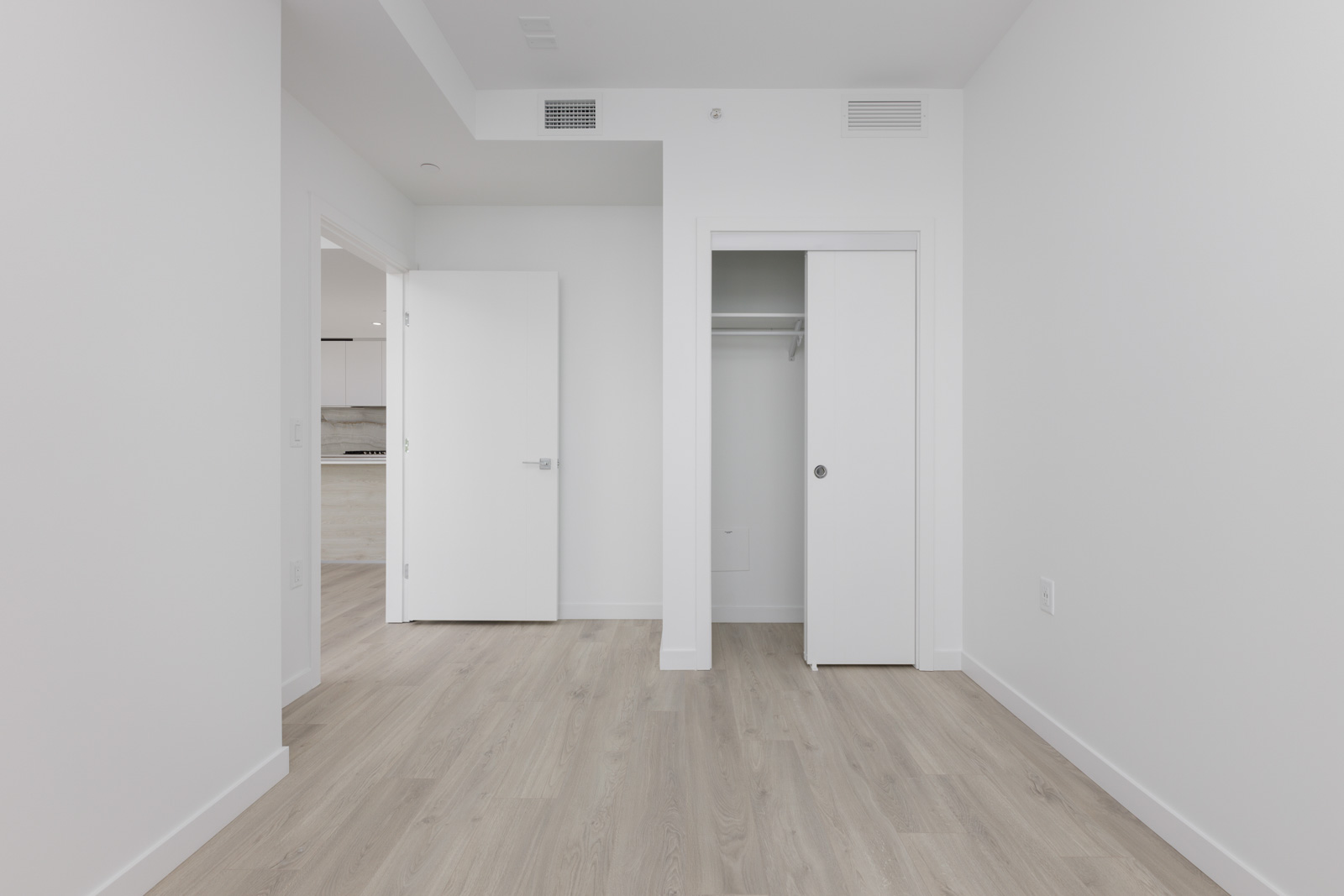 Empty, unfurnished room with light wood flooring, white walls, an open door leading to another area, and a small closet with sliding doors.