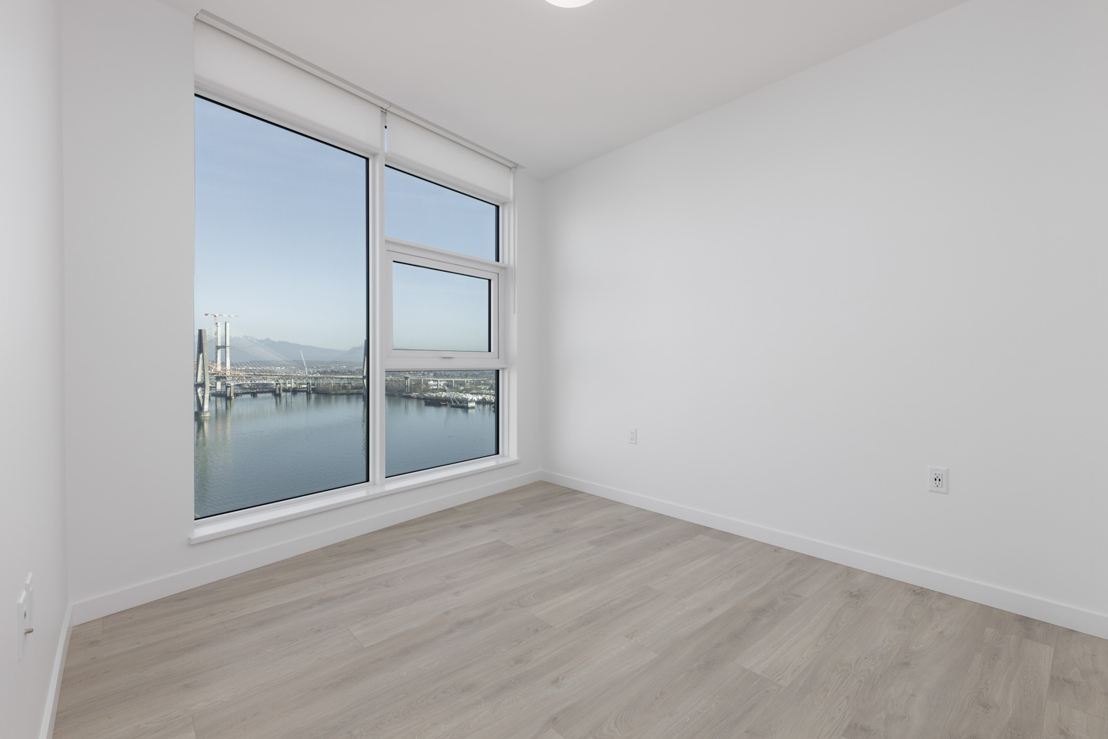 Empty room with light wood flooring, white walls, and a large window overlooking a body of water and a bridge construction site.