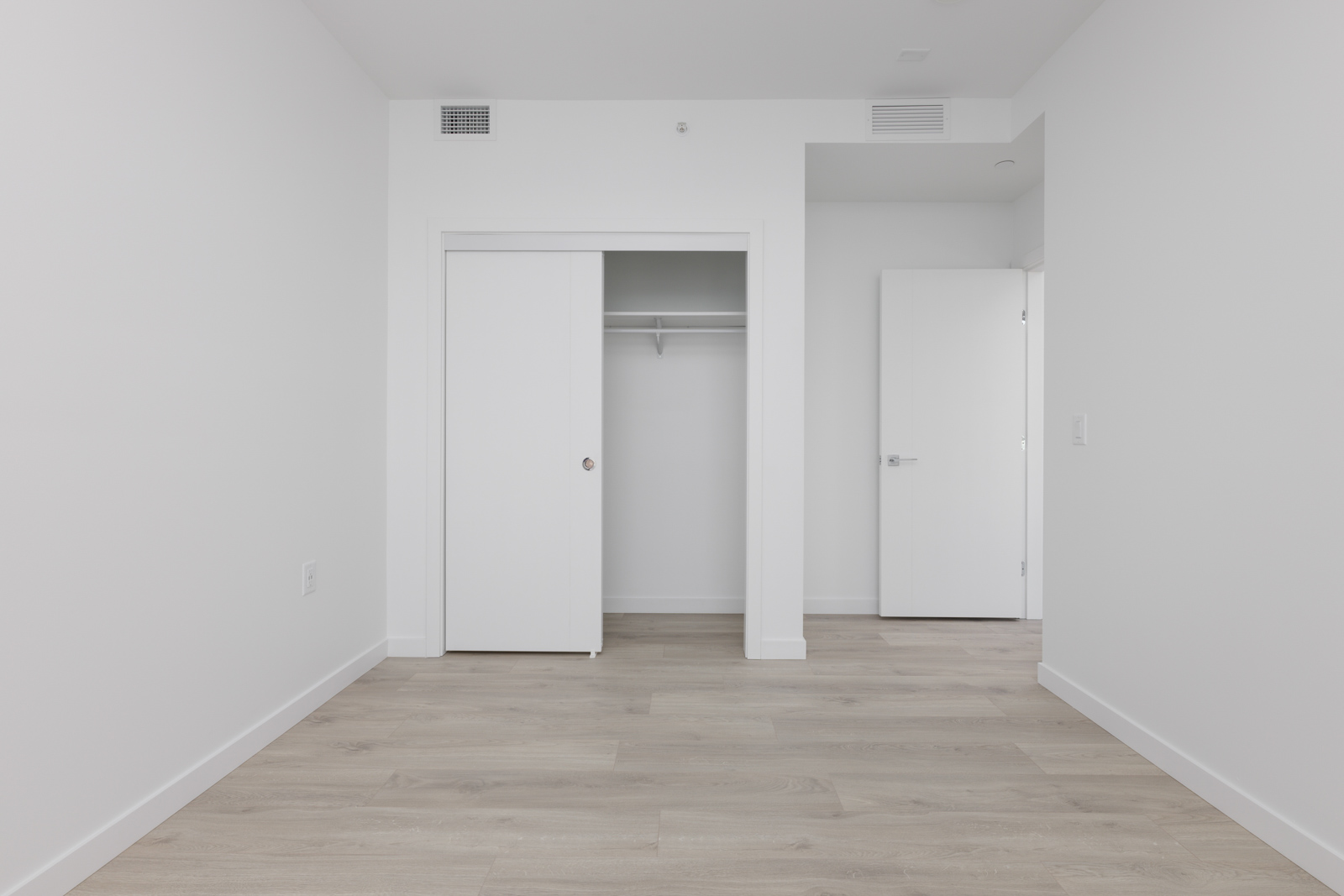 Empty room with light wood flooring, white walls, an open closet with sliding doors, and an open door leading to another space.