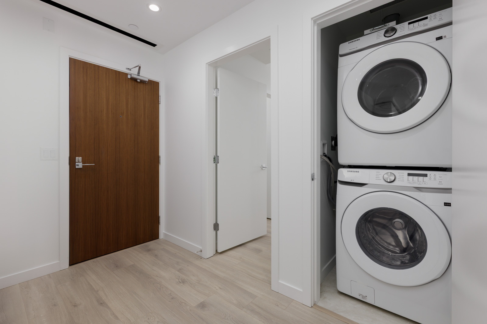 A stacked washer and dryer unit is installed in a closet next to a hallway with a wooden door and light-colored flooring.