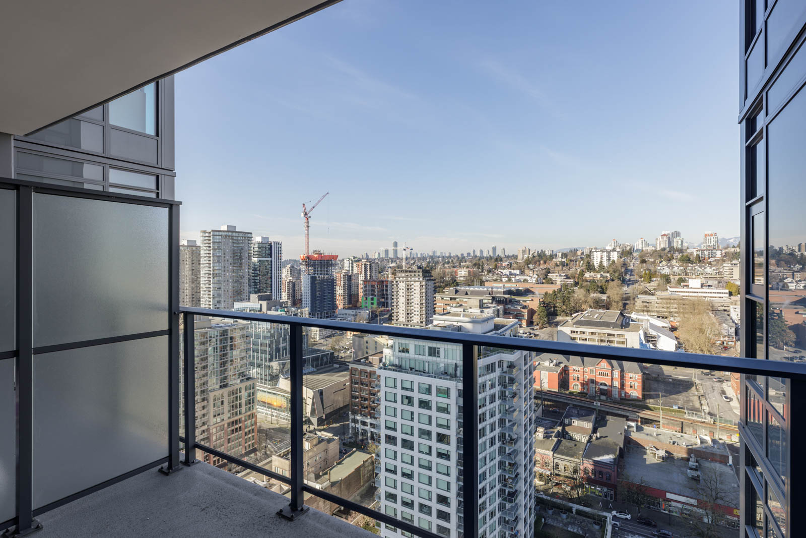 View from a high-rise balcony overlooking a cityscape with modern buildings, a construction crane, streets, and a clear sky in the background.