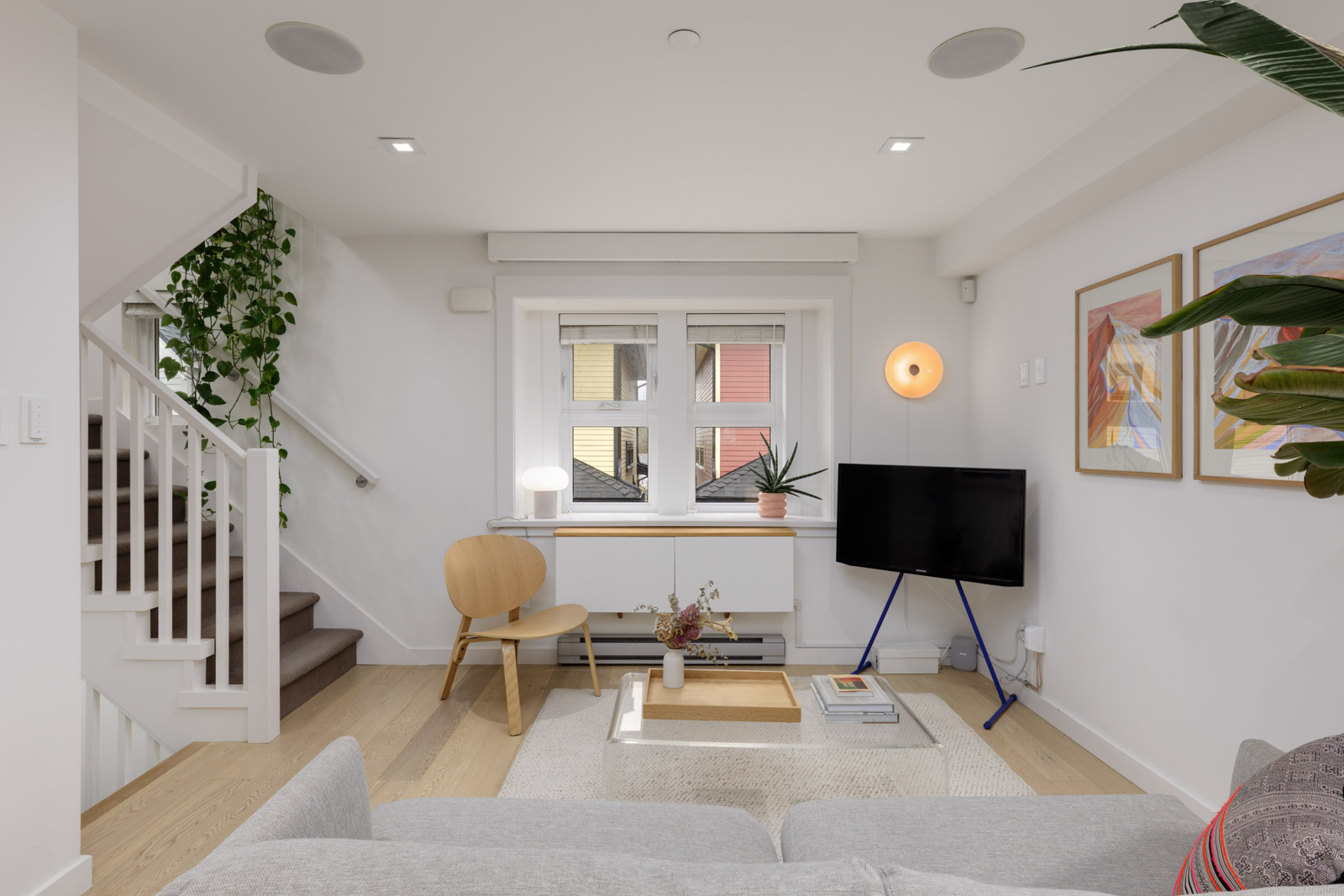 Modern living room with light wood floors, neutral furniture, a small round table with a chair, a TV on a tripod stand, wall art, houseplants, and stairs to the left.