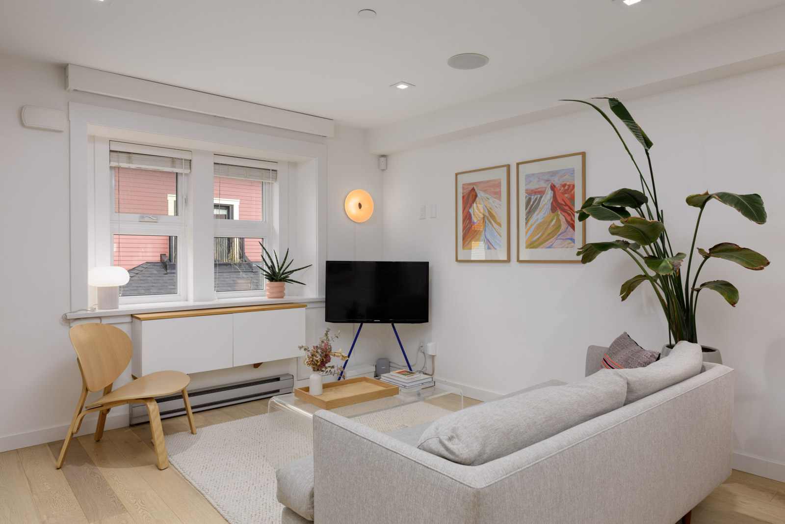 A modern living room with a light gray sofa, flat-screen TV, wooden chair, large potted plant, and wall art; natural light enters through a window with a pink building visible outside.