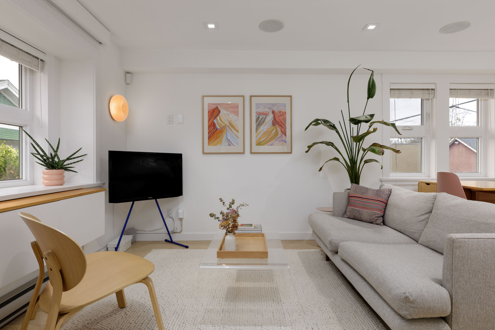 Modern living room with a light gray sofa, wooden chair, small TV, potted plants, coffee table, and two abstract art prints on the white wall. Natural light fills the space.