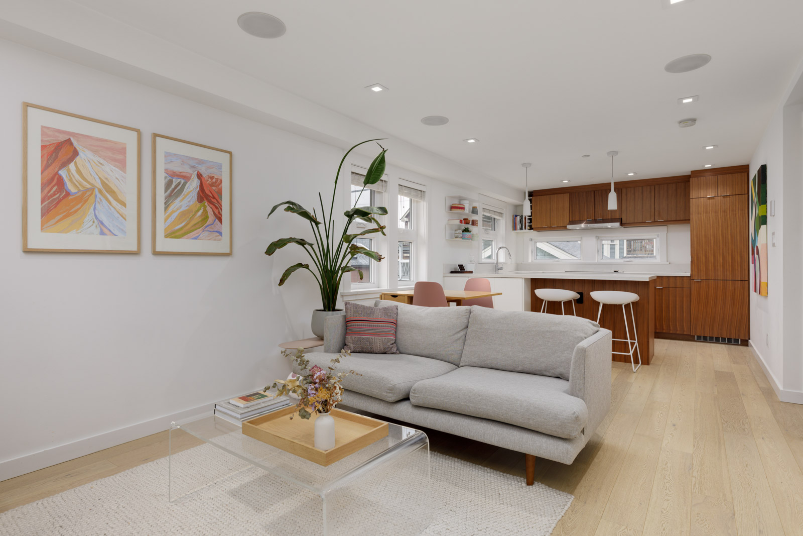 Modern open-plan living room and kitchen with light wood floors, a gray sofa, large plant, wall art, and white cabinetry with wooden accents.