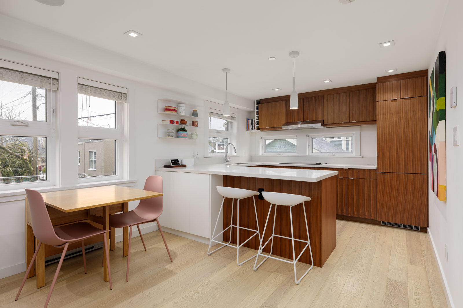 Modern kitchen with wood cabinets, white countertops, island with two white bar stools, dining table with pink chairs, open shelves, and large windows letting in natural light.