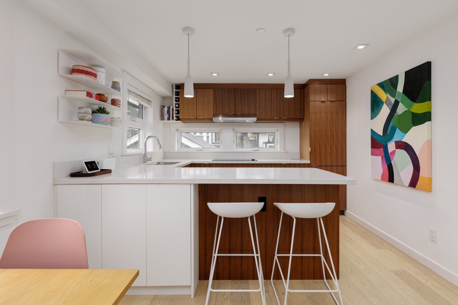Modern kitchen with white countertops, wooden cabinets, two white bar stools, open shelves, and a colorful abstract painting on the wall.