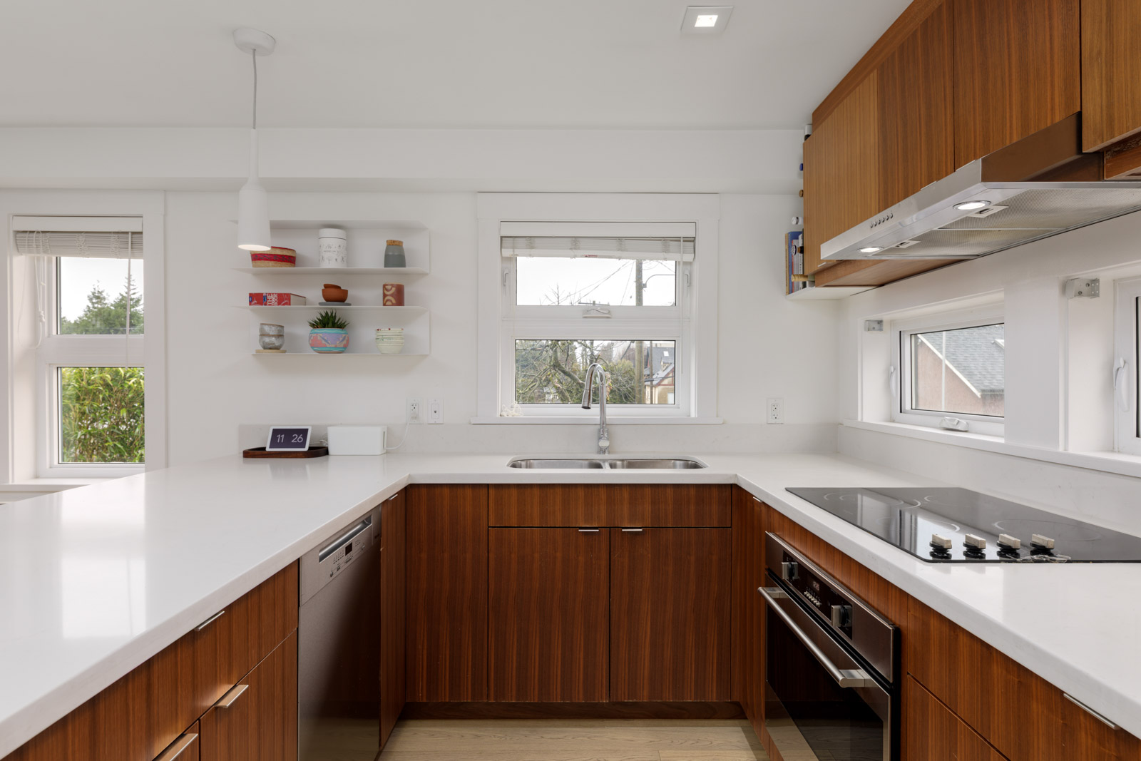 Modern kitchen with white countertops, wooden cabinets, electric cooktop, built-in oven, sink under a window, and open shelves with decor items on the wall.