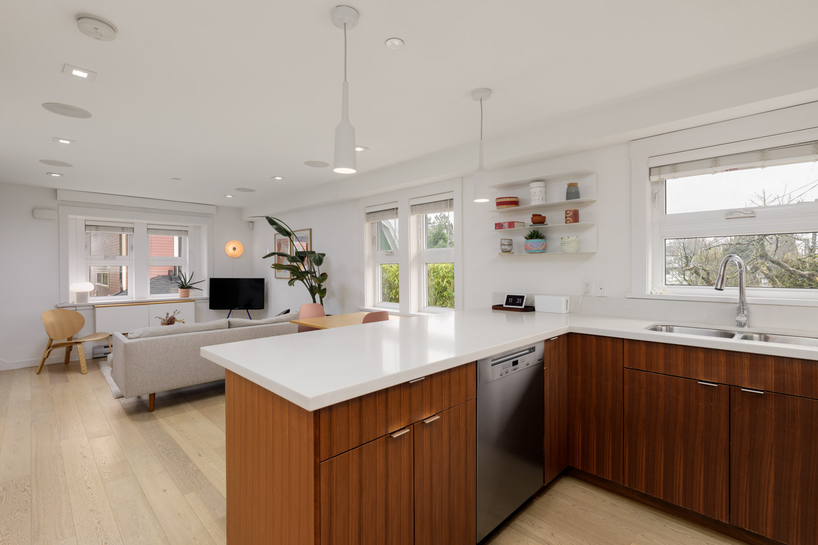 Modern open-plan kitchen and living area with white walls, wood cabinets, light wood flooring, and large windows letting in natural light.
