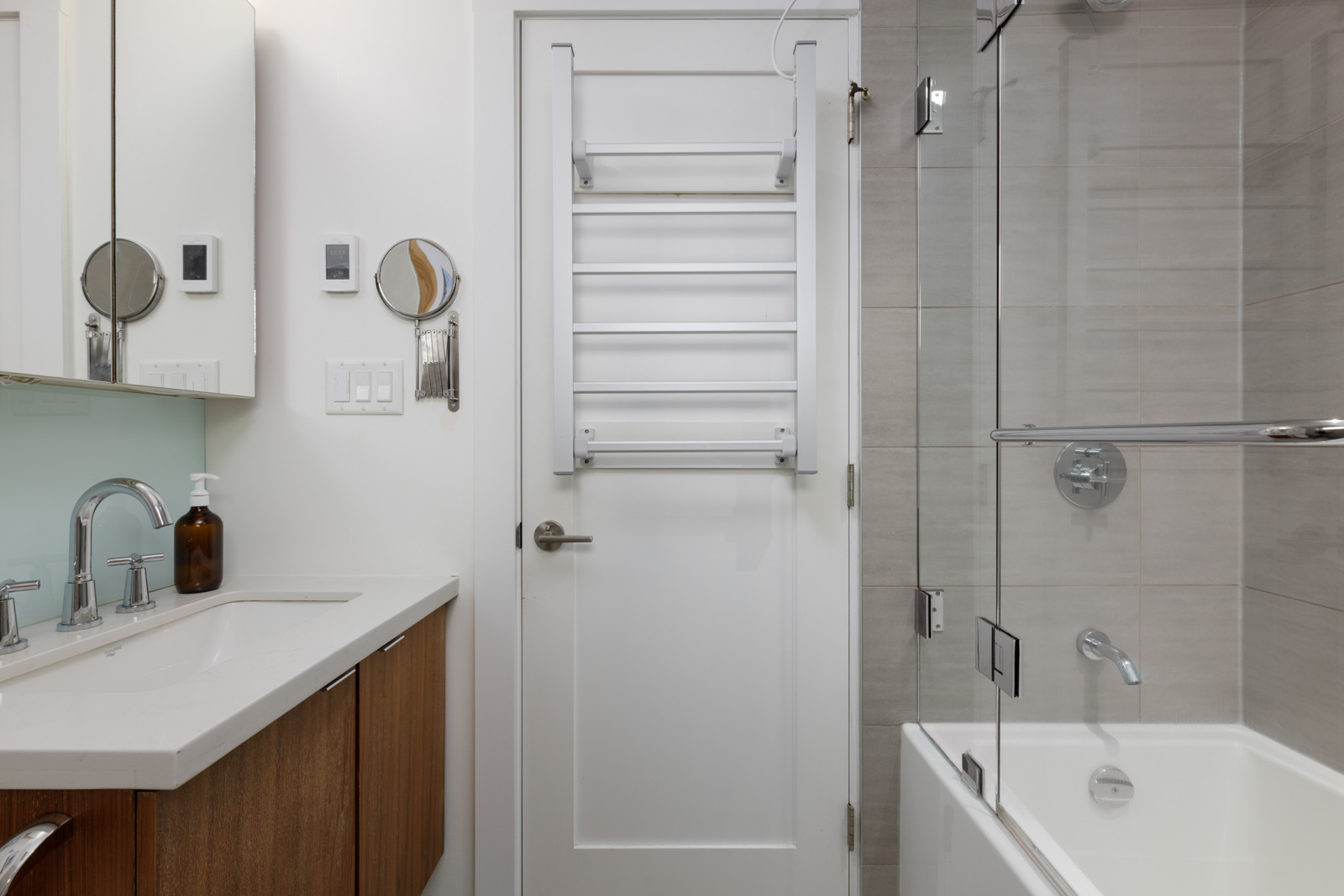 Modern bathroom with a white door, towel rack, glass shower enclosure, bathtub, and vanity with a sink and mirror.
