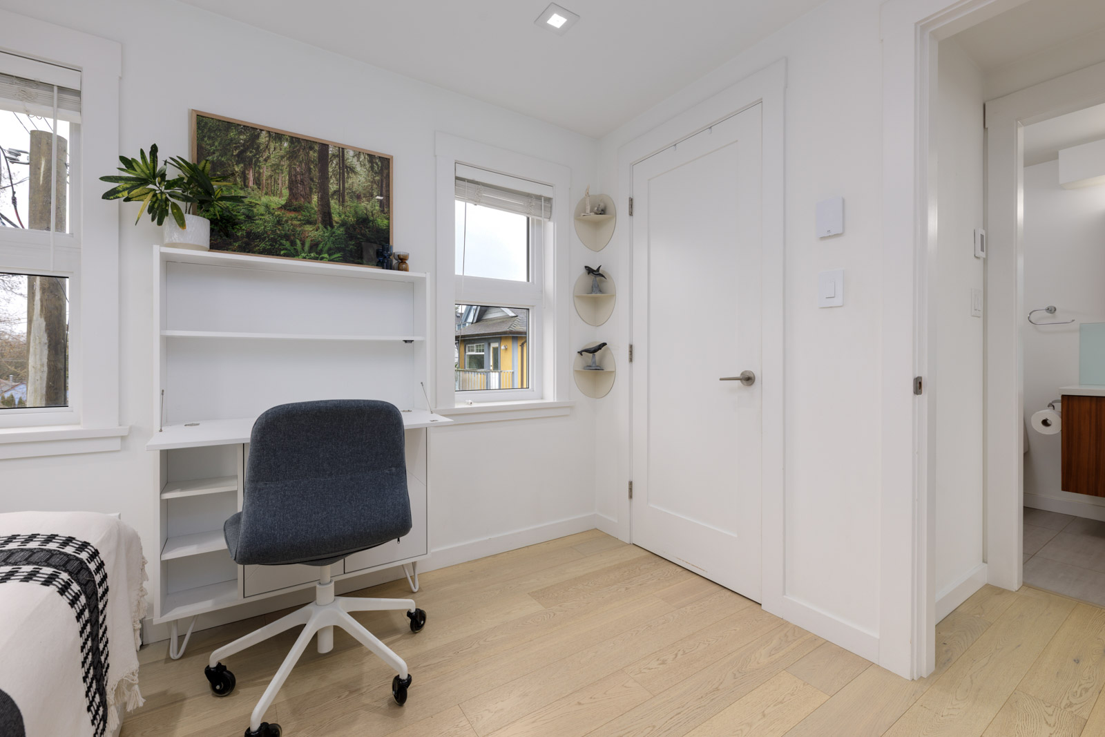 A minimalist home office with a white desk, gray office chair, wall shelves, and a framed forest photo, located next to a bedroom and bathroom.