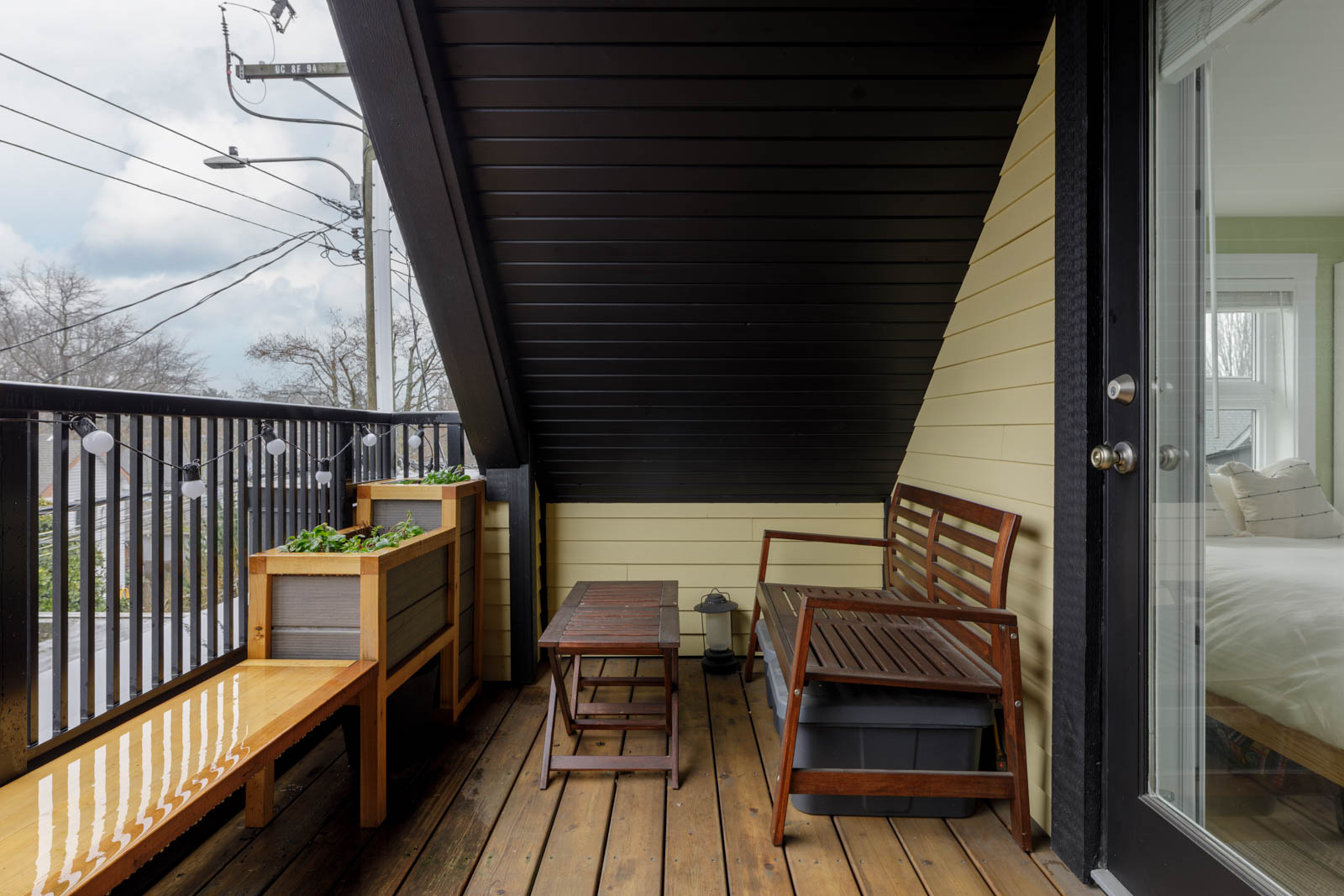 A small covered balcony with wooden floor, bench seating, planters, a coffee table, black railing, and a view of power lines and overcast sky. A glass door leads to a bedroom.