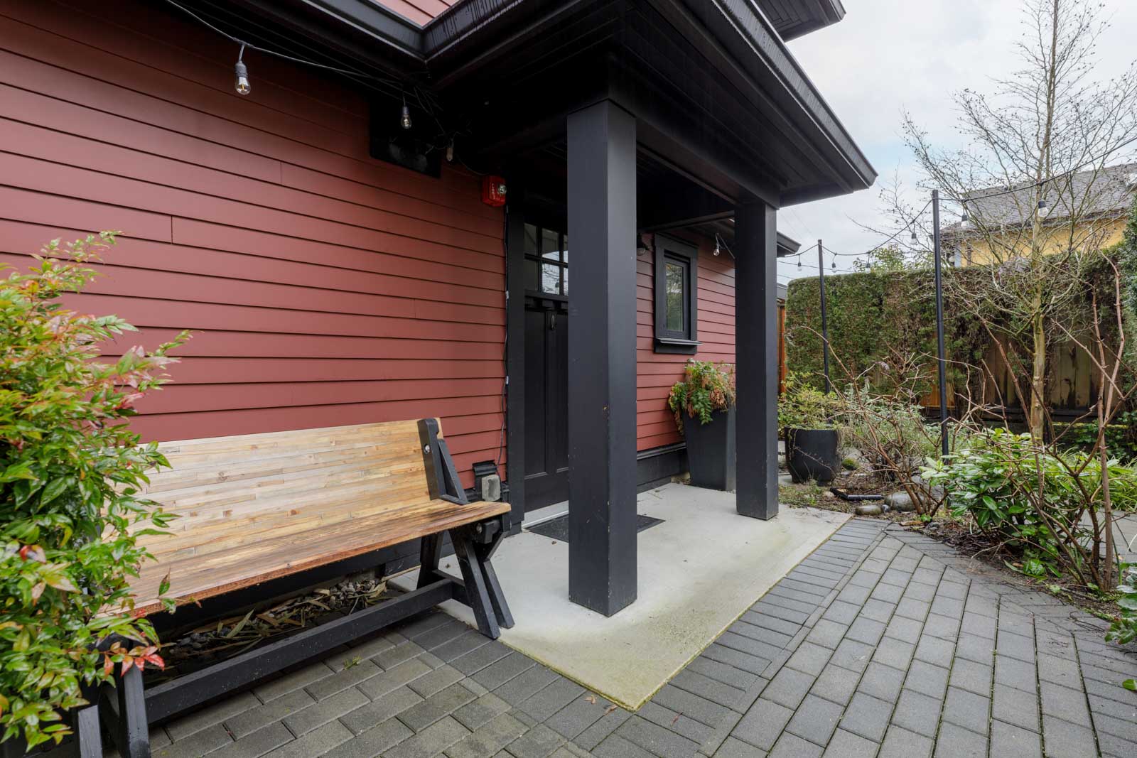 Wooden bench beside the entrance of a red house with black trim, supported by two black columns, surrounded by greenery and paved walkway.