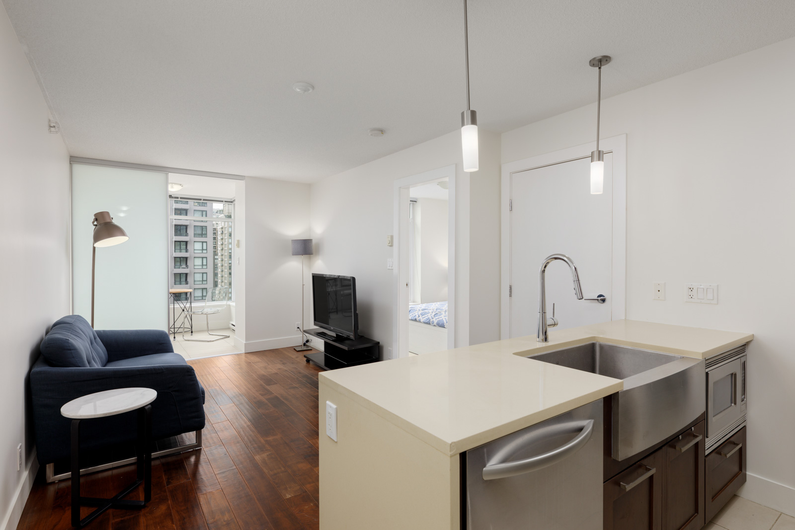 Modern apartment interior with a kitchen island, stainless steel sink, living area with a sofa and TV, hardwood floors, and a view of a city building through large windows.