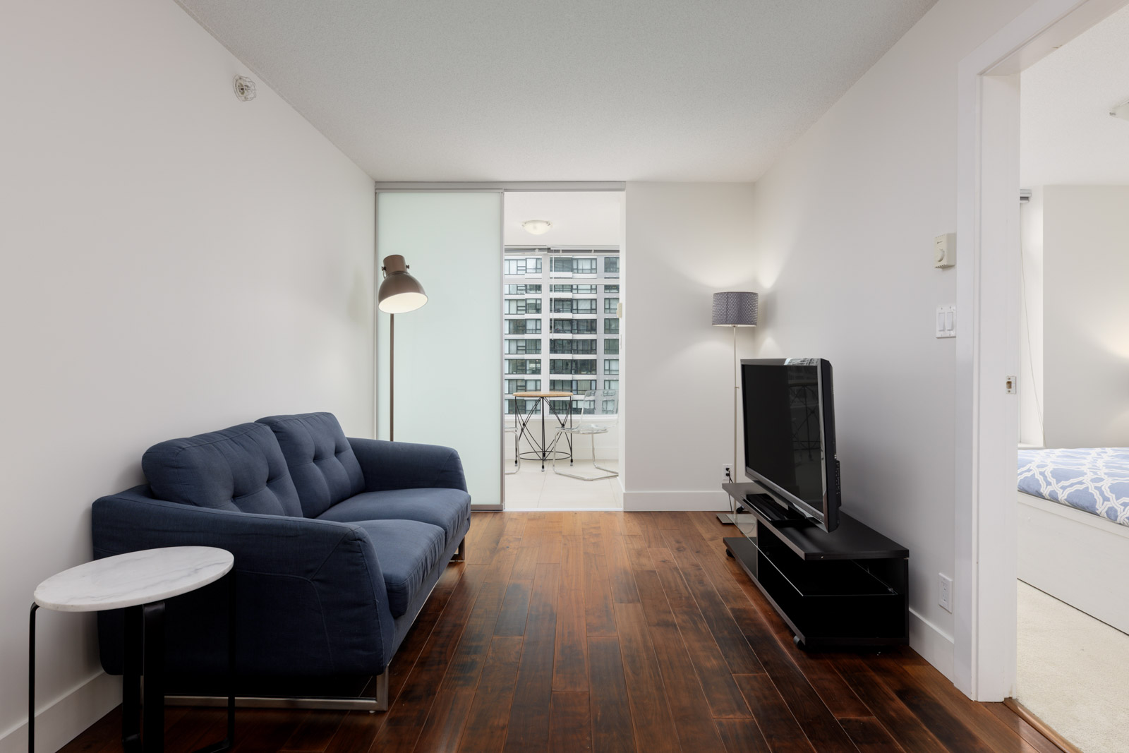 Modern living room with a blue sofa, floor lamp, TV on a stand, round side table, and hardwood floors, leading to a bright dining area with large windows.