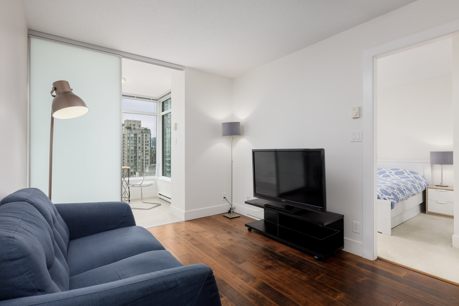Modern living room with a blue sofa, floor lamp, TV on a stand, and hardwood floors. Bedroom and balcony with city view are visible through open doors.