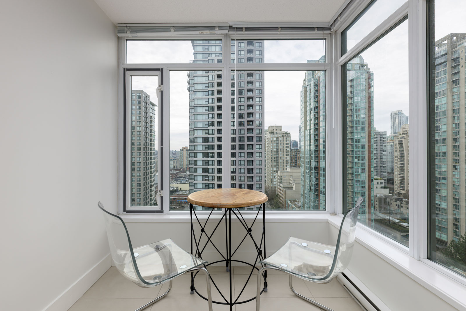 A small round table with two clear chairs is set by large windows overlooking tall city buildings on a cloudy day.