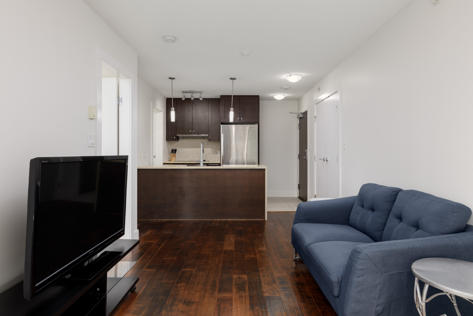 Modern apartment living room with a blue sofa, flat-screen TV on a stand, and a view of a kitchen with dark cabinets and stainless steel appliances in the background.