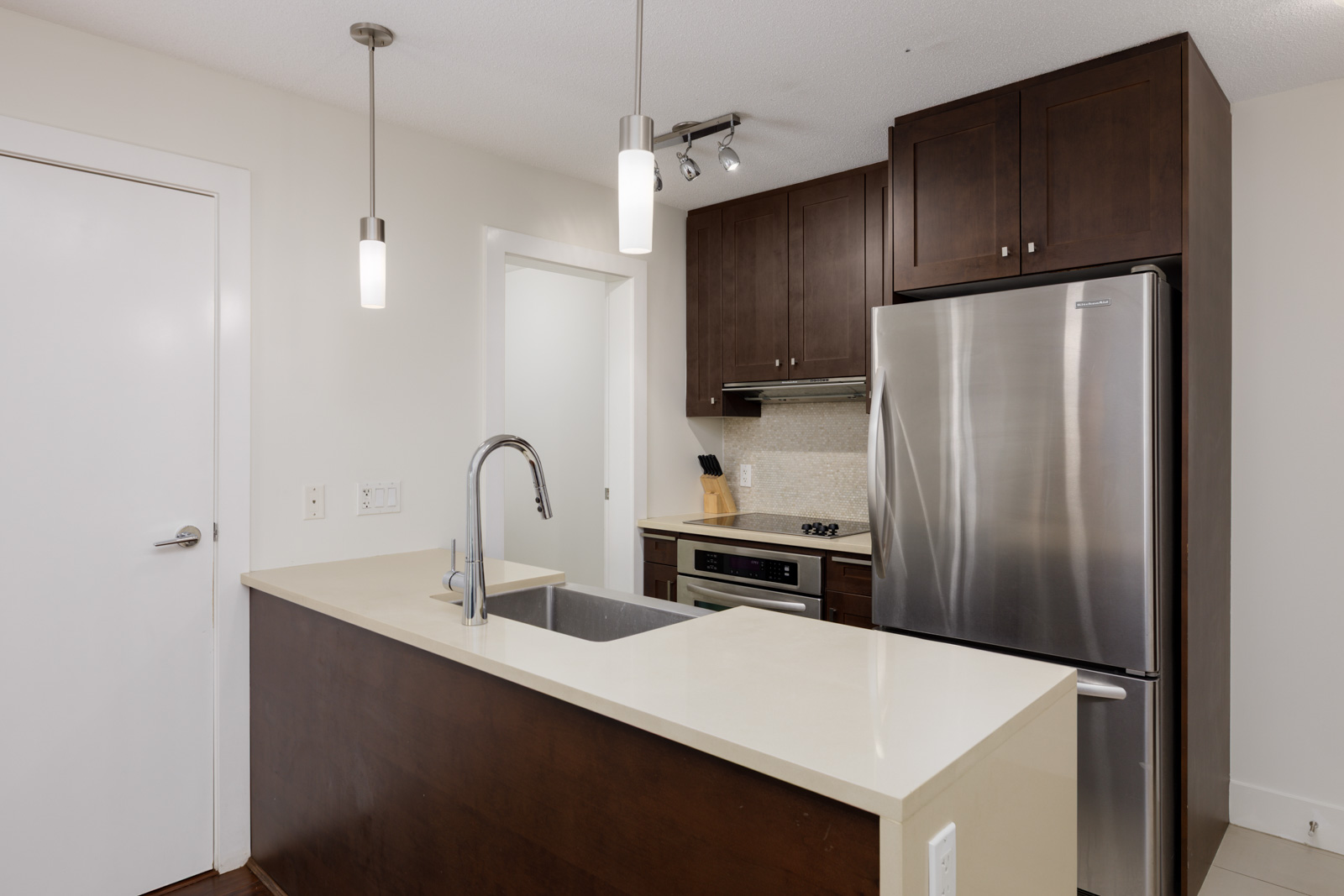 Modern kitchen with dark wood cabinets, stainless steel refrigerator, built-in oven, electric cooktop, and a white countertop island with sink and hanging pendant lights.