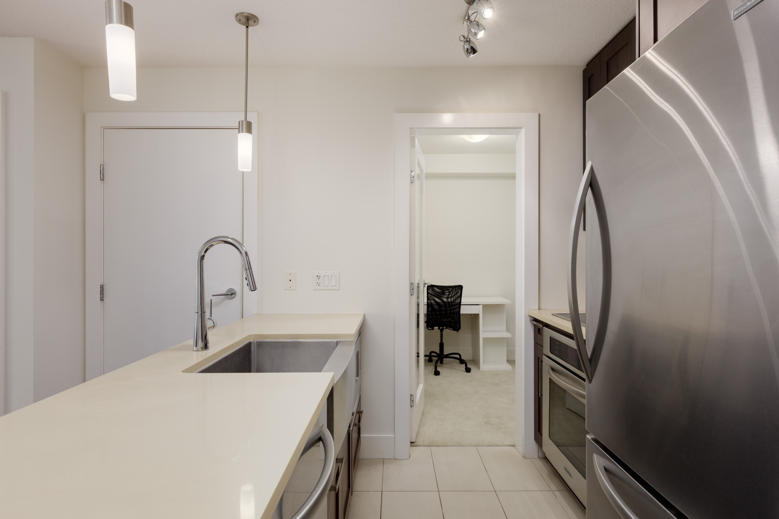 Modern kitchen with stainless steel appliances, a sink island, and a view into a small office space with a desk and chair through an open doorway.