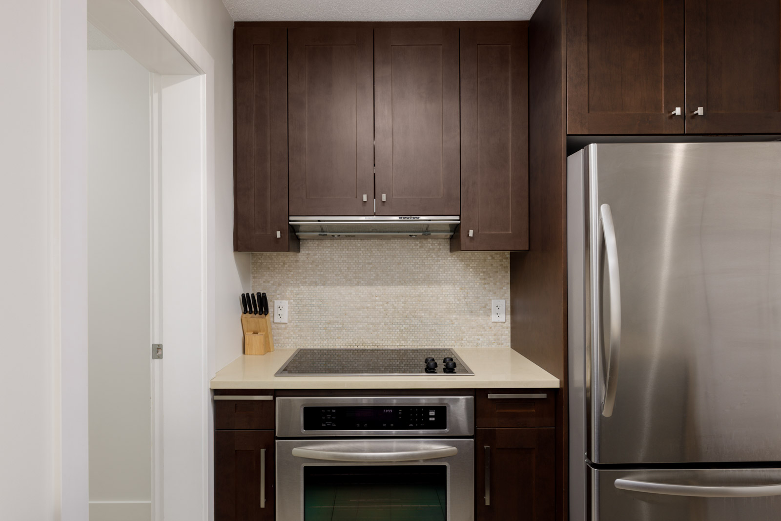 Modern kitchen with dark wood cabinets, stainless steel oven and refrigerator, electric cooktop, and a knife block on a light countertop against a tile backsplash.