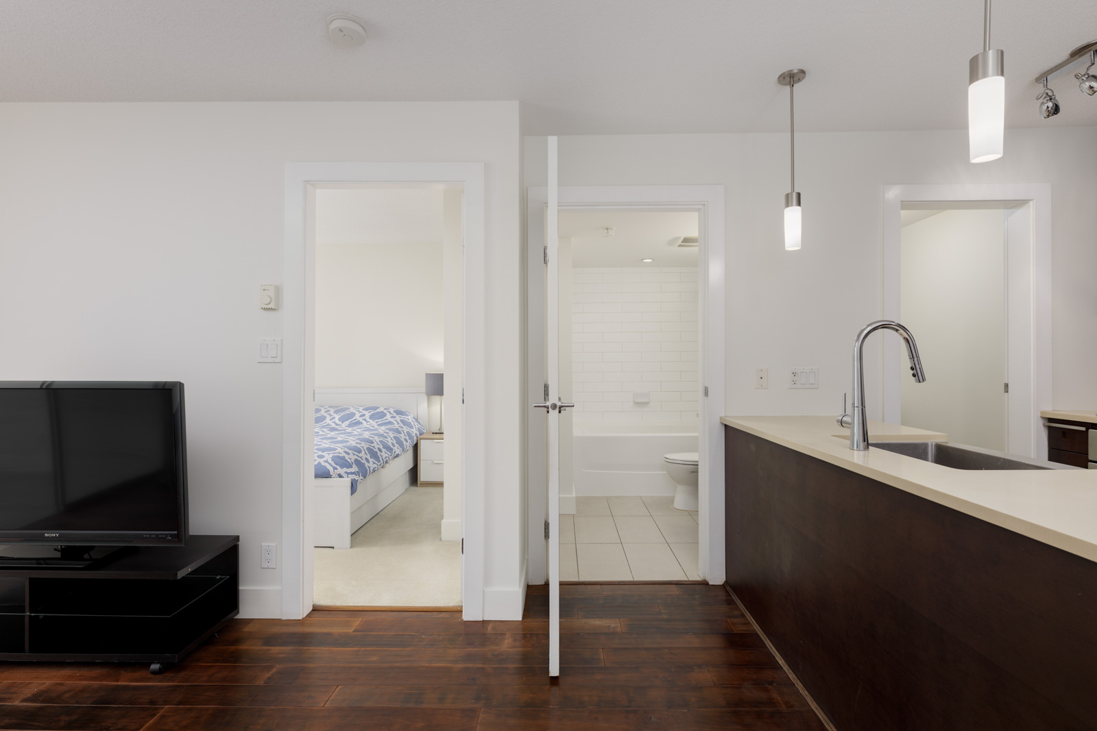 Modern apartment interior showing a living area with TV, open kitchen with hanging lights, and doorways leading to a bedroom and a bathroom with white tiles.