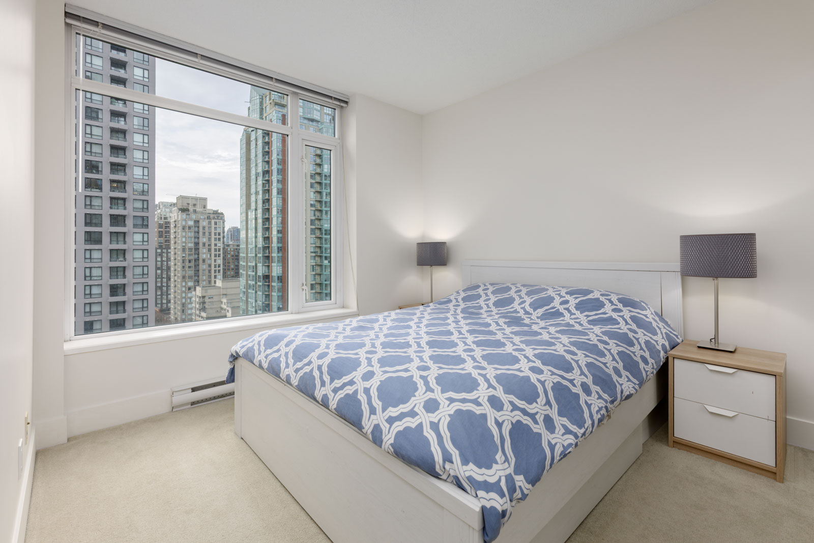 Modern bedroom with a large window showing city skyscrapers, a double bed with blue patterned bedding, two lamps, and a white nightstand.