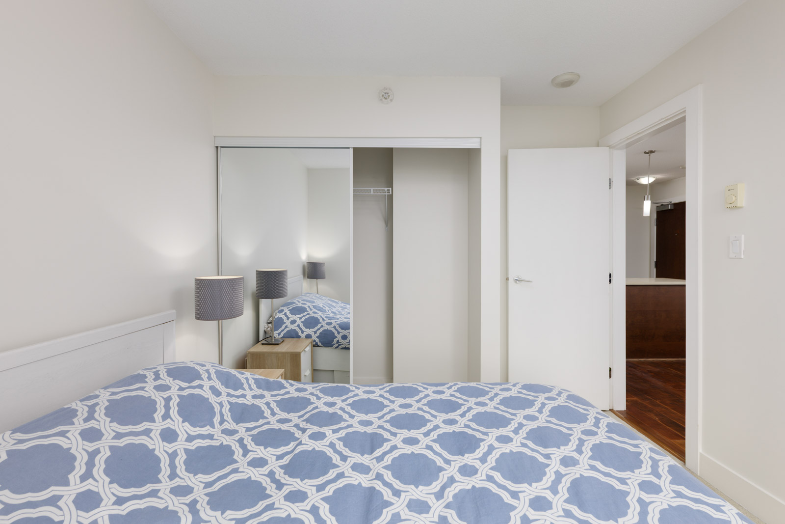 A simple bedroom with a bed covered in a blue and white patterned duvet, a mirrored closet, a nightstand with a lamp, and an open door leading to a hallway.