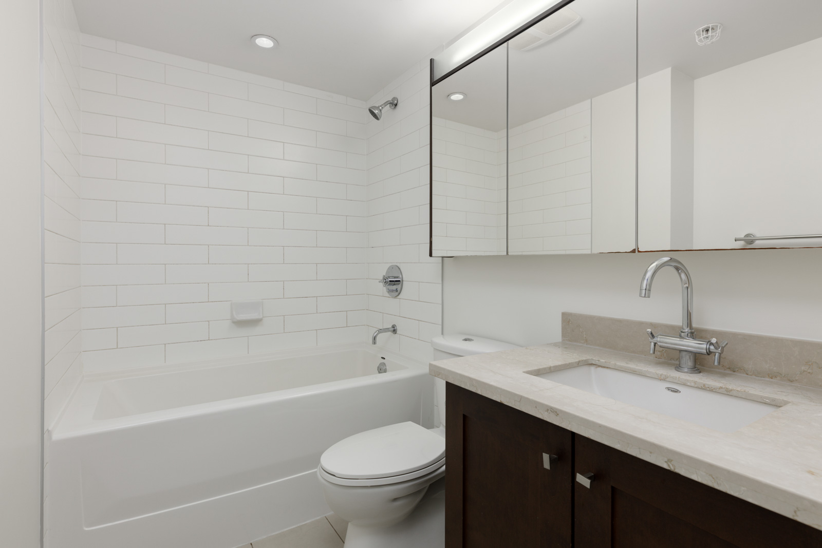Modern bathroom with white tile walls, a bathtub-shower combination, toilet, and a vanity with a sink, faucet, and mirrored medicine cabinet.