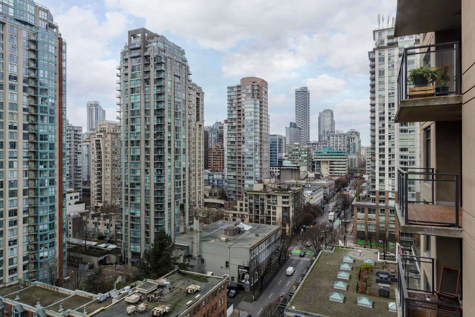 View of a cityscape with multiple high-rise buildings, a busy street below, and overcast sky. Some balconies and rooftop structures are visible in the foreground.