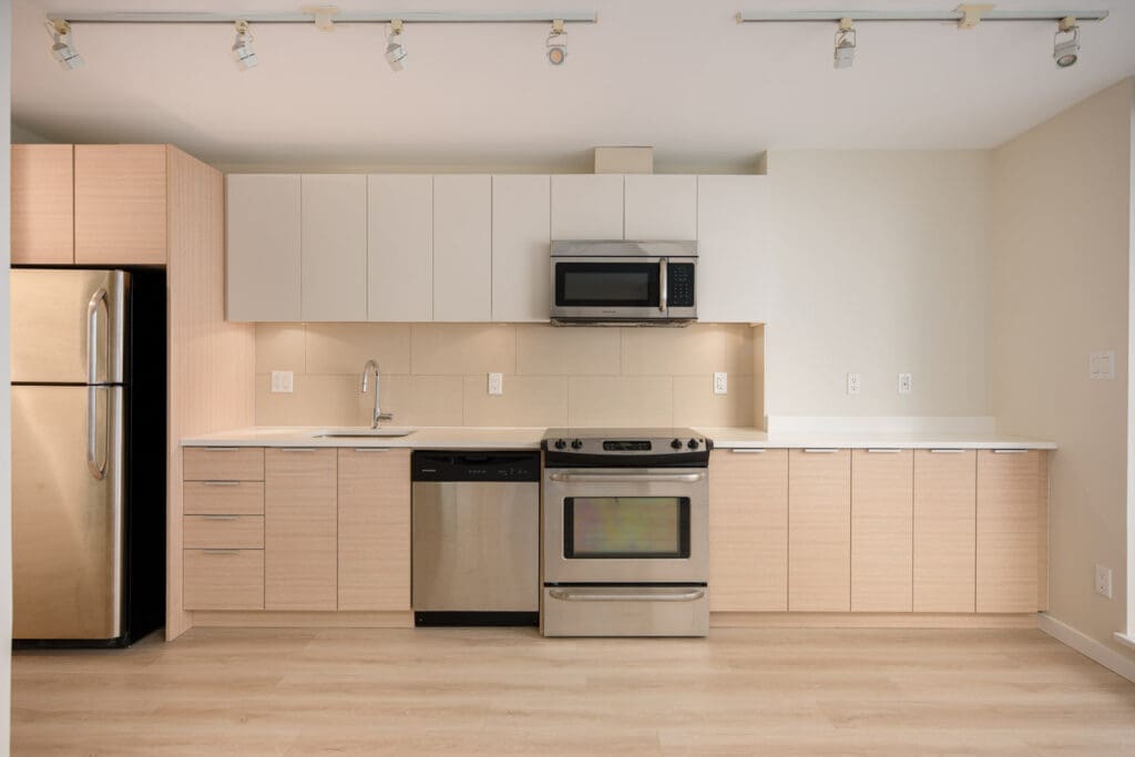 Modern kitchen with light wood cabinets, stainless steel refrigerator, dishwasher, oven, and microwave, under track lighting on a light wood floor.