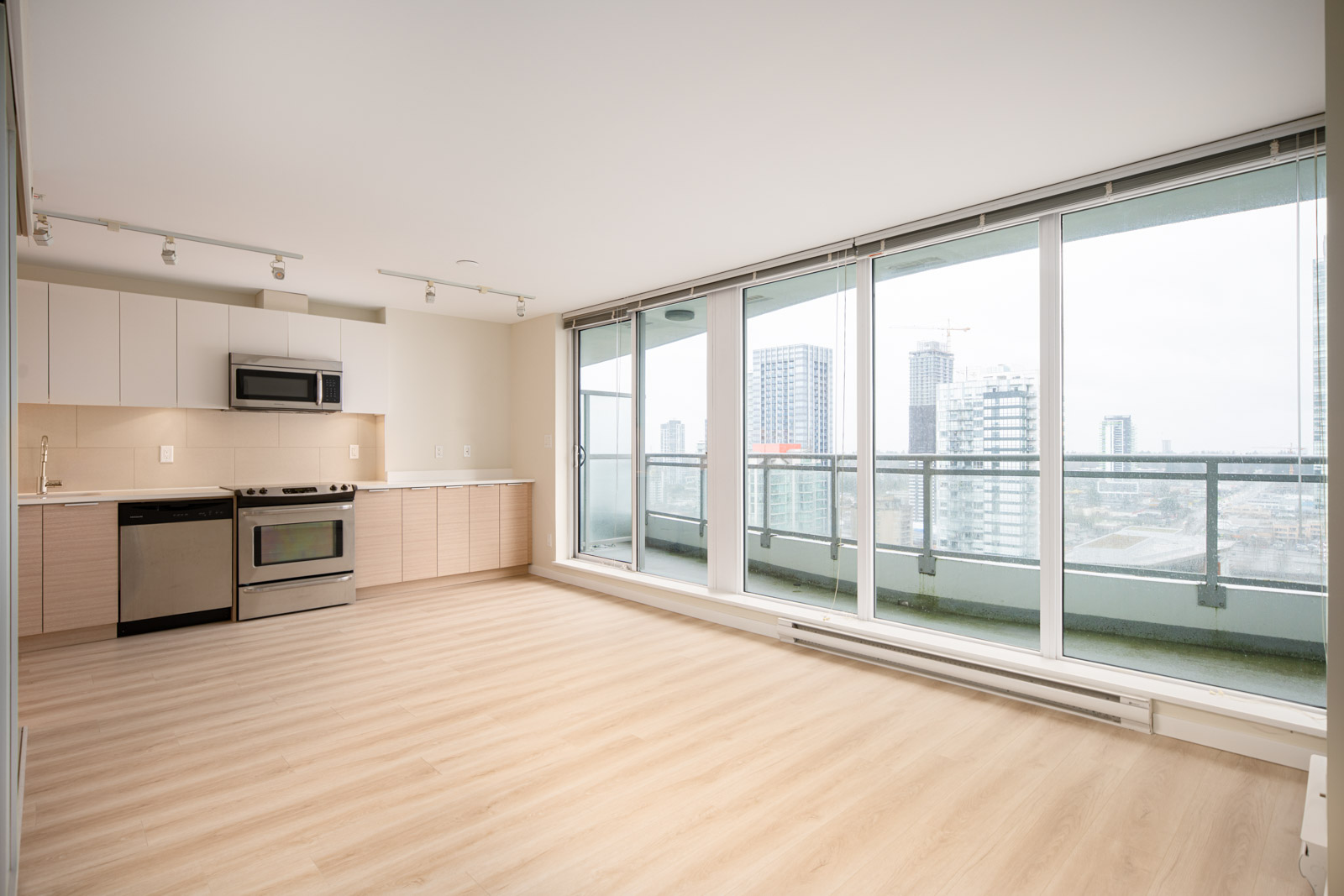 Unfurnished modern apartment with wood flooring, open kitchen, stainless steel appliances, and large floor-to-ceiling windows overlooking a city skyline and balcony.